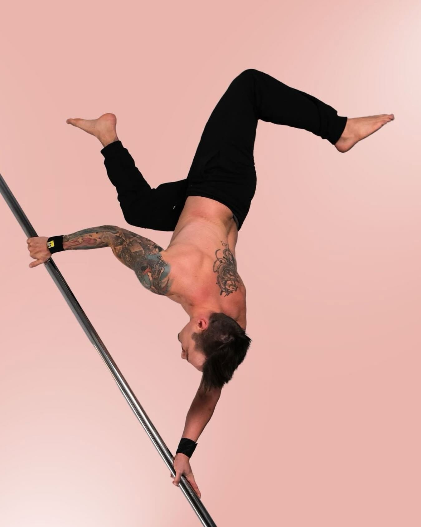 A shirtless, tattooed male pole athlete holding himself upside down on a slanted pole with both hands, wearing black SneakyDeez wrist wraps against a soft pink background