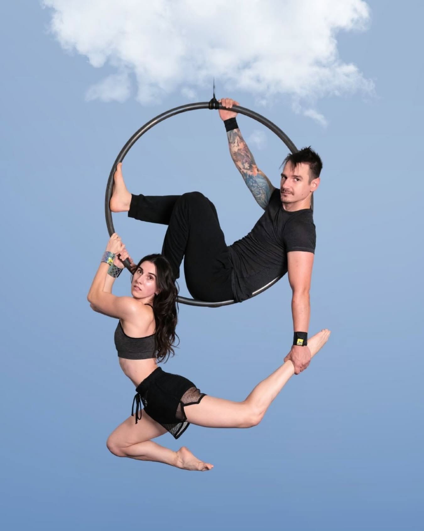 Two aerialists on an aerial hoop against a blue sky backdrop, a man sitting in the hoop and a woman hanging below it with one hand on the hoop, both wearing SneakyDeez wrist wraps