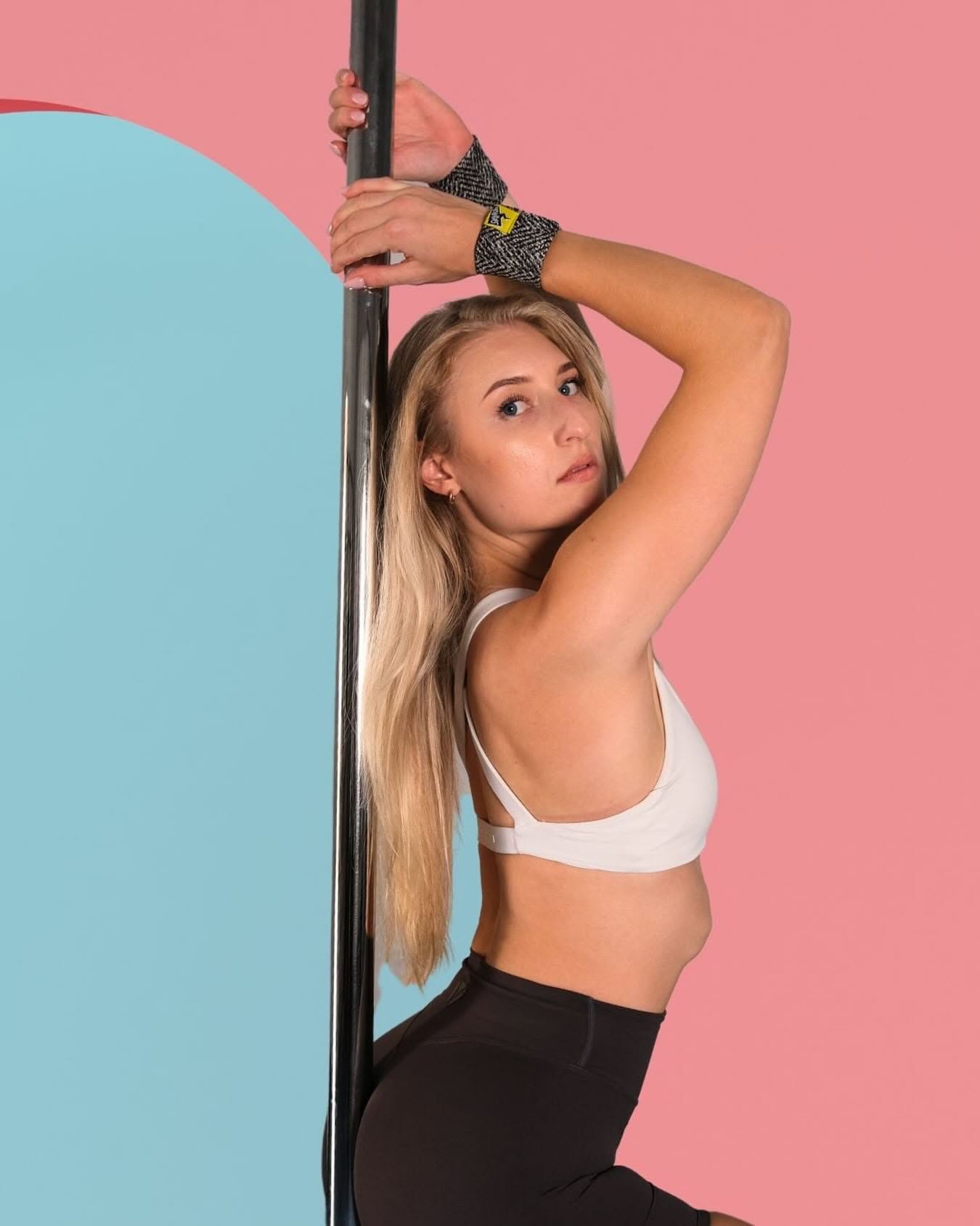 A pole dancer with long blonde hair holding the pole above her head, wearing grey patterned SneakyDeez wrist wraps against a pink and powder-blue backdrop