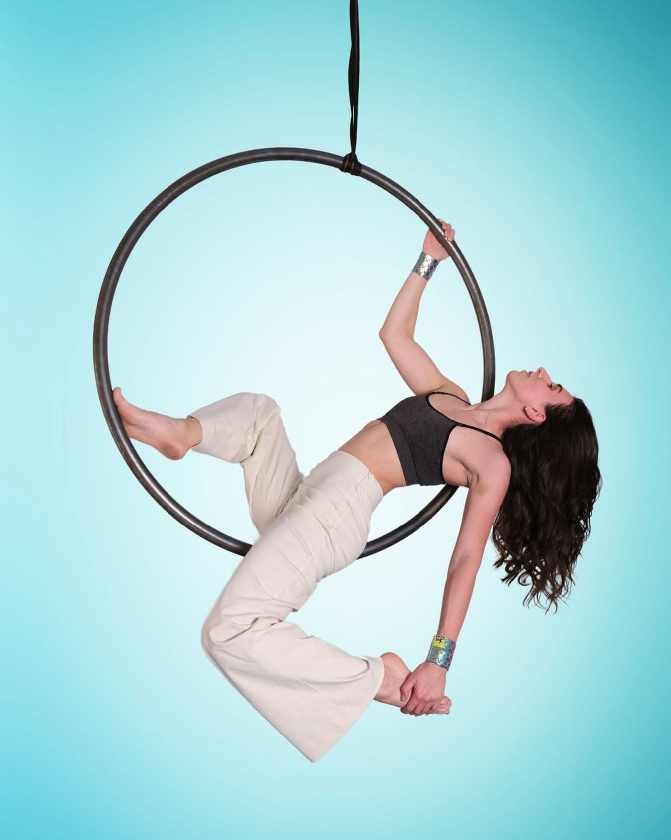 An aerial hoop artist in white trousers and a dark crop top leaning back inside the hoop and holding the bar with one hand, wearing SneakyDeez wrist wraps