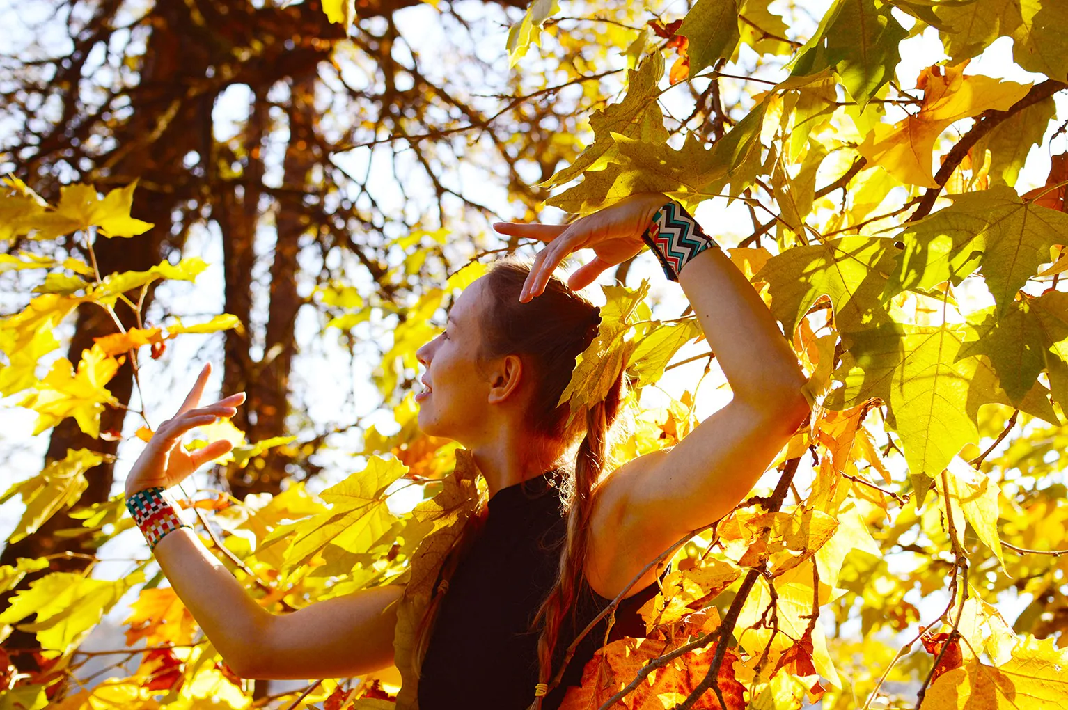 A woman with a braid dancing among autumn leaves with her arms raised, wearing colourful patterned wrist wraps on both wrists
