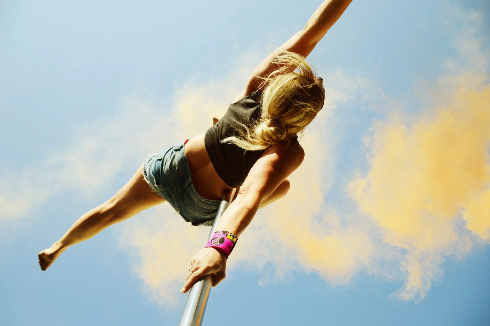 A pole dancer photographed from below against a blue sky, holding herself out from the pole with one hand and wearing a pink wrist wrap on that wrist