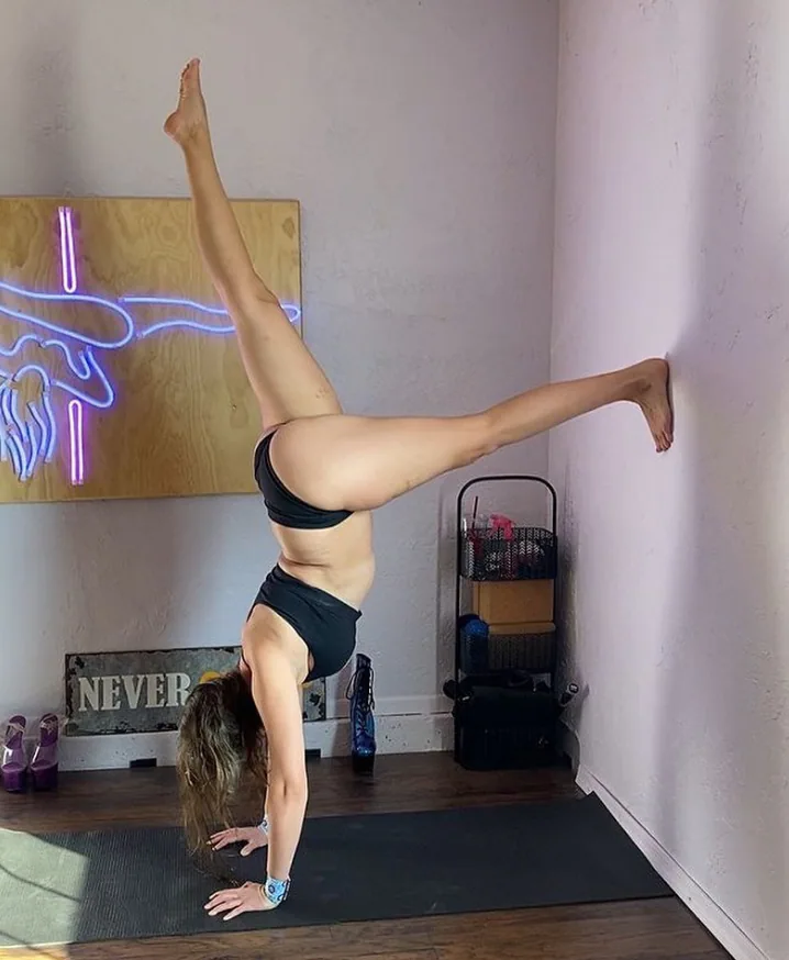 A SneakyDeez customer in a handstand against the wall of her home studio, with one foot on the wall, her hands on a dark mat and a wrap on her wrist