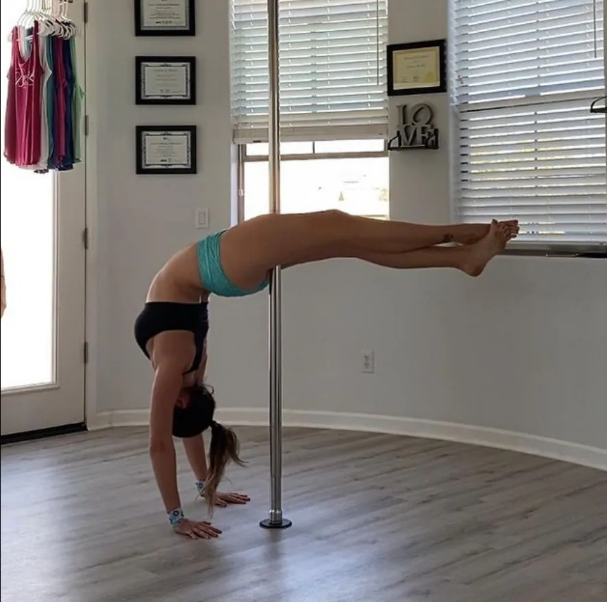 A SneakyDeez customer balancing on her hands beside a pole in a bright studio, with her hips against the pole and her legs stretched out level behind her
