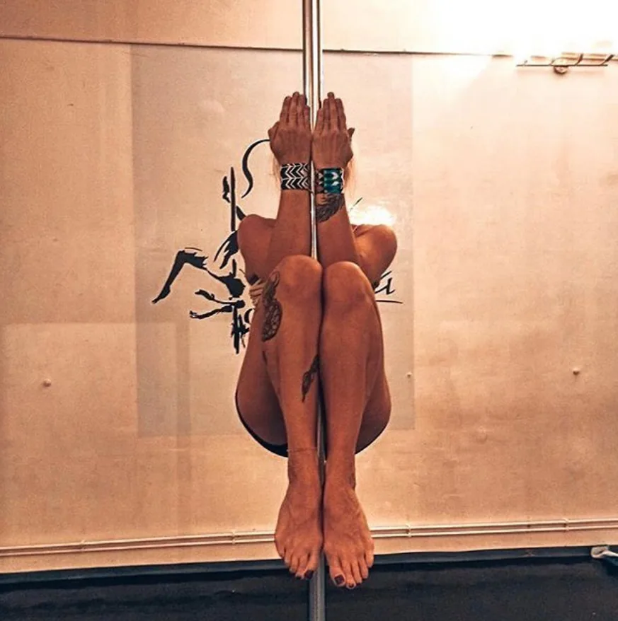 A SneakyDeez customer curled up in a tucked hold on the pole with her knees to her chest and her face hidden, her hands pressed against the pole above her head and patterned wrist wraps on both wrists