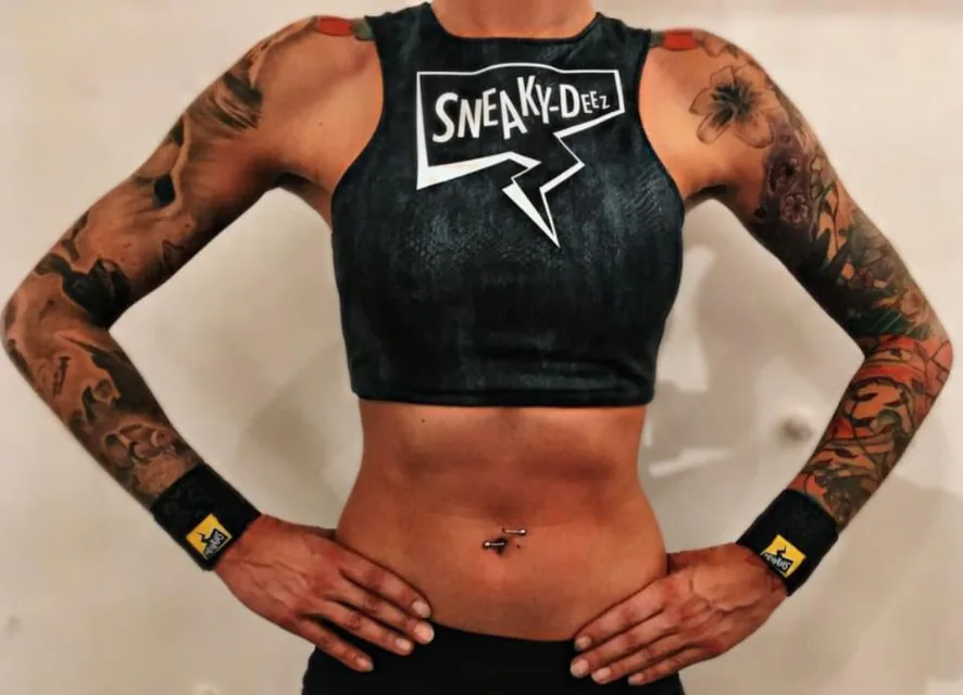 A woman with tattooed arms in a black SneakyDeez crop top standing with her hands on her hips, wearing black wrist wraps that leave her thumbs and fingers free