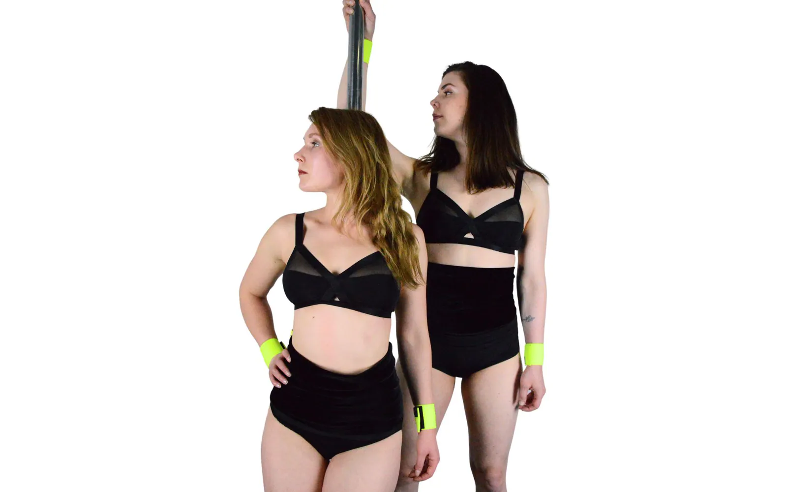 Two dancers in black bra tops standing by a pole, both wearing black waist warmers over high-waisted shorts and neon yellow wrist wraps