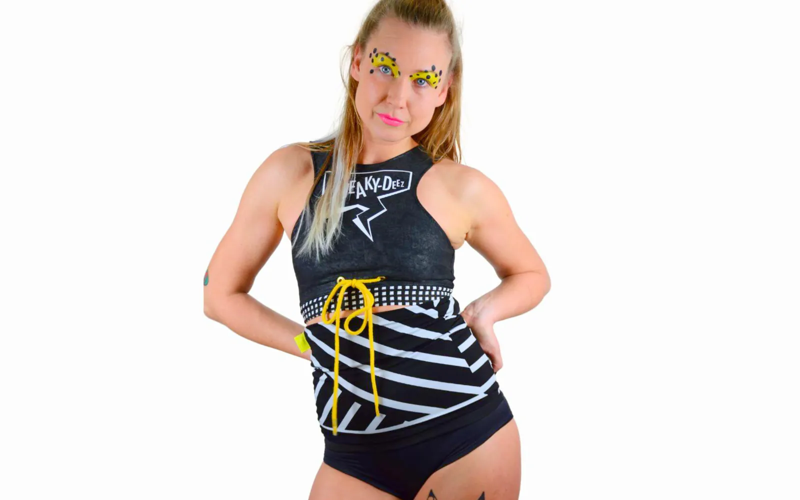 Woman in a black SneakyDeez crop top wearing a black and white striped waist warmer laced at the front with a yellow cord, with yellow and pink face paint